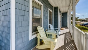 Une véranda avec deux fauteuils Adirondack et une petite table, un espace couvert avec une rambarde, et une vue sur un parking et une rue en arrière-plan.