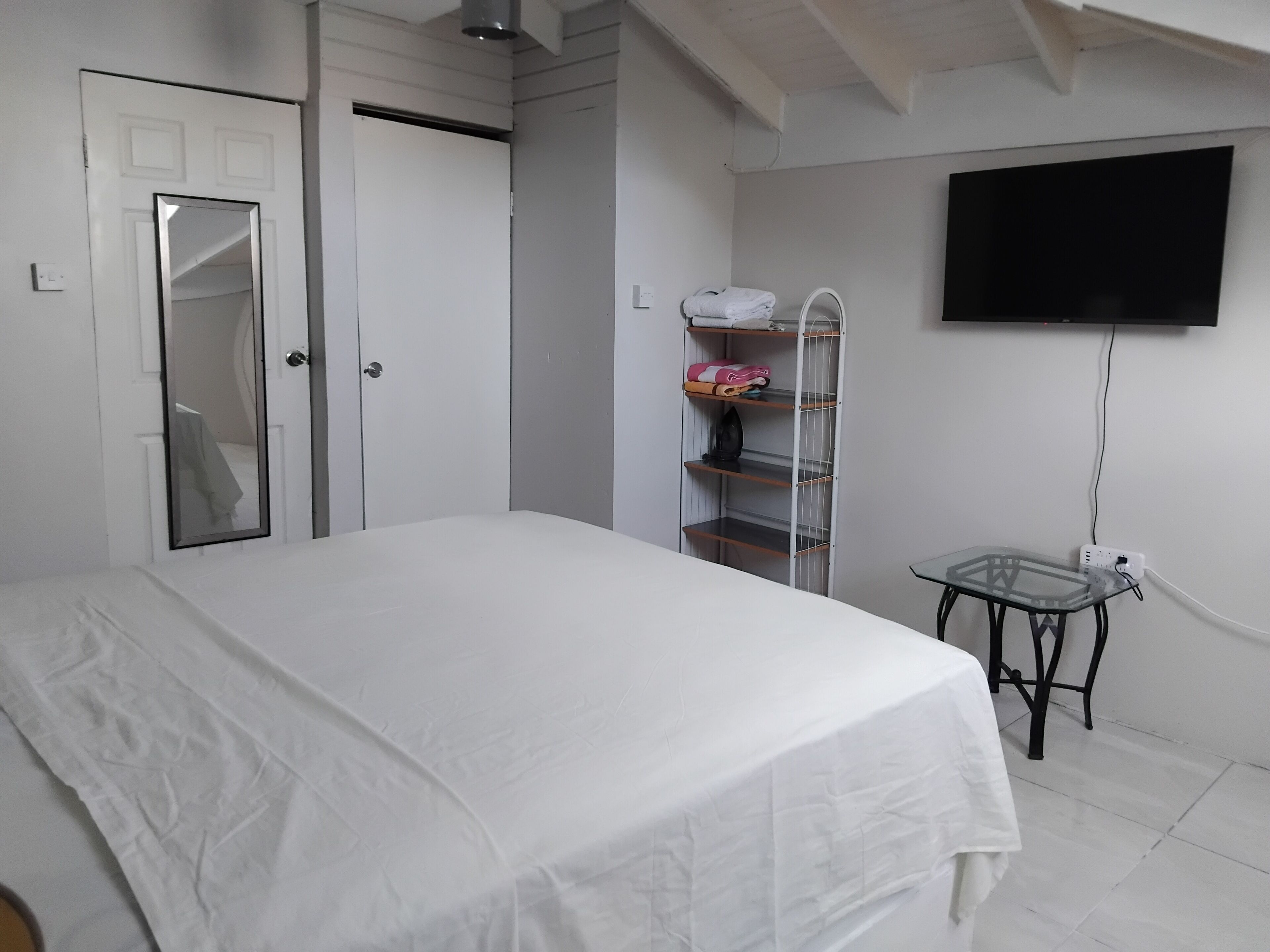Un dormitorio con cama, armario, espejo, estante con toallas dobladas, una mesita y televisor montado en la pared.