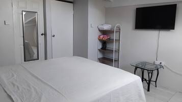 Un dormitorio con cama, armario, espejo, estante con toallas dobladas, una mesita y televisor montado en la pared.