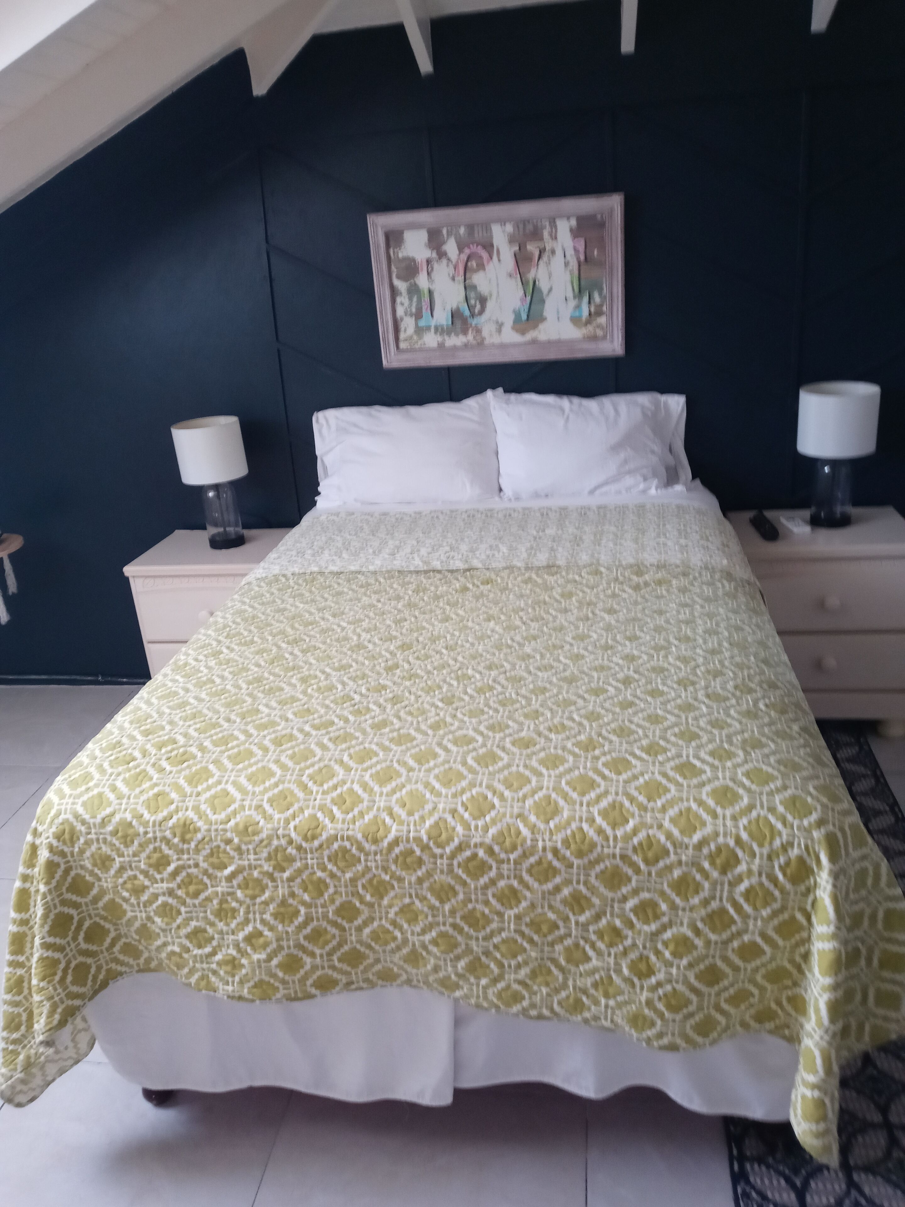 A bedroom with a bed, two bedside tables, a nightstand, a wall-mounted artwork, and a ceiling with a skylight.