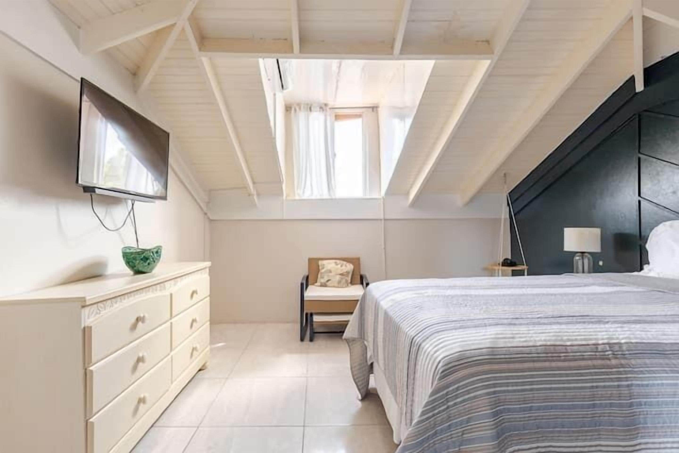 A bedroom with a sloped ceiling, a bed with a striped bedspread, a dresser with drawers, a chair with a cushion, a bedside table with a lamp, and a television mounted on the wall.