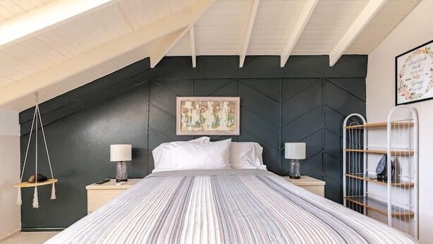 Un dormitorio con techo inclinado, una cama con ropa de cama a rayas, dos mesitas de noche con lámparas, un sillón colgante y un estante fijado a la pared.