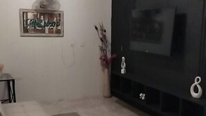 Una sala de estar con un televisor de pantalla plana grande montado en una pared oscura, una mesita con un florero y una zona de estar con cojines.