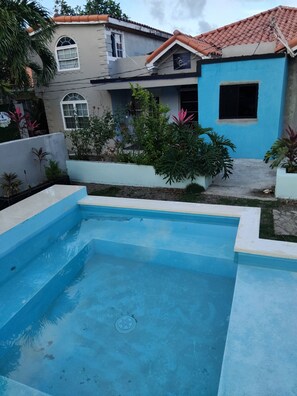 A swimming pool with a blue interior, surrounded by a garden with various plants and a building with a tiled roof in the background. - Leisure Duplex At Csompo Silver Manor (Tower Isle)