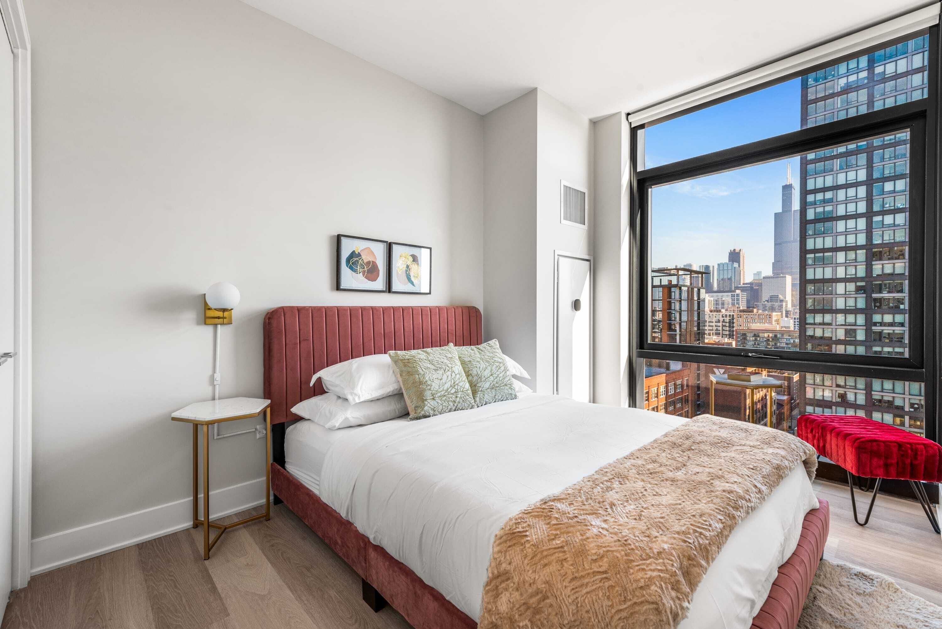 A modern bedroom with a large bed, a red headboard, and a city view through a large window.