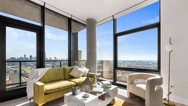 A modern living room with a sofa, coffee table, and armchair, large windows with a city view, and a tall column. - City Views + Walk to Dining, Ideal for All (Chicago)