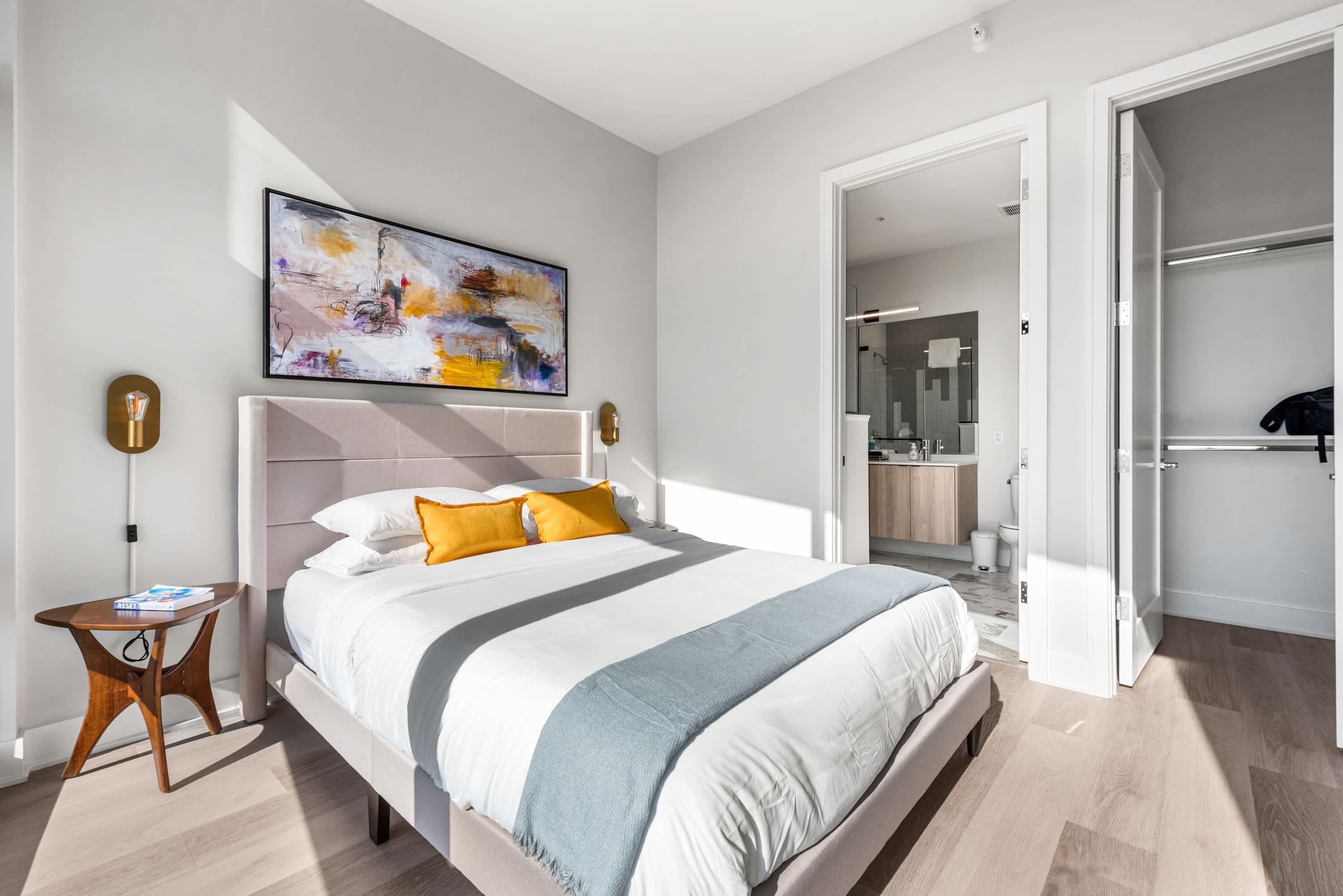 A modern bedroom with a large bed, a bedside table, a painting, and a bathroom visible through an open door.