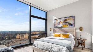 A modern bedroom with a large window, a bed with white and gray bedding, a colorful abstract painting, and a small wooden side table. - City Views + Walk to Dining, Ideal for All (Chicago)