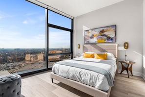 A modern bedroom with a large window, a bed with white and gray bedding, a colorful abstract painting, and a small wooden side table.