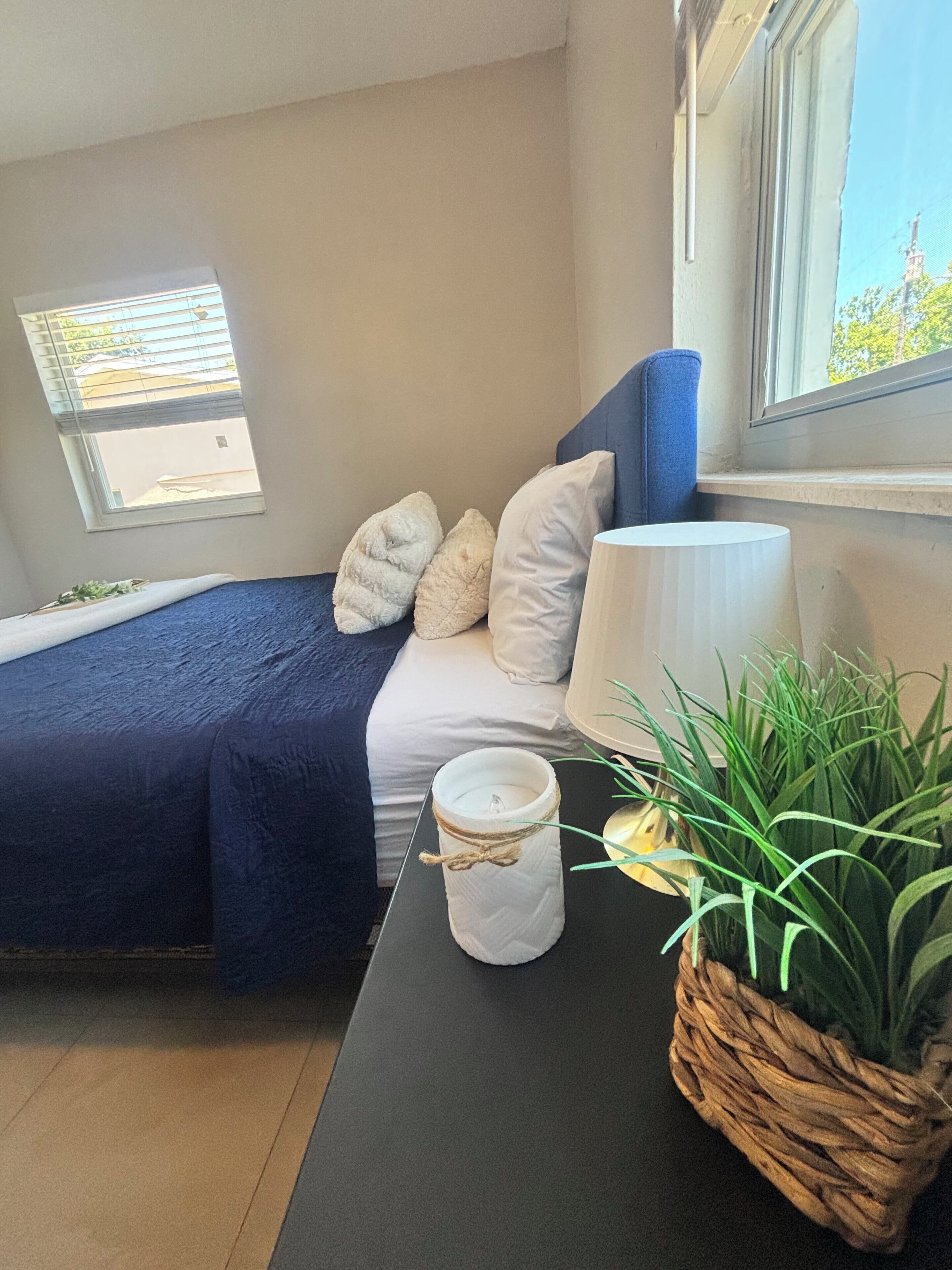 A bedroom with a bed, pillows, a bedside lamp, a window, and a plant.