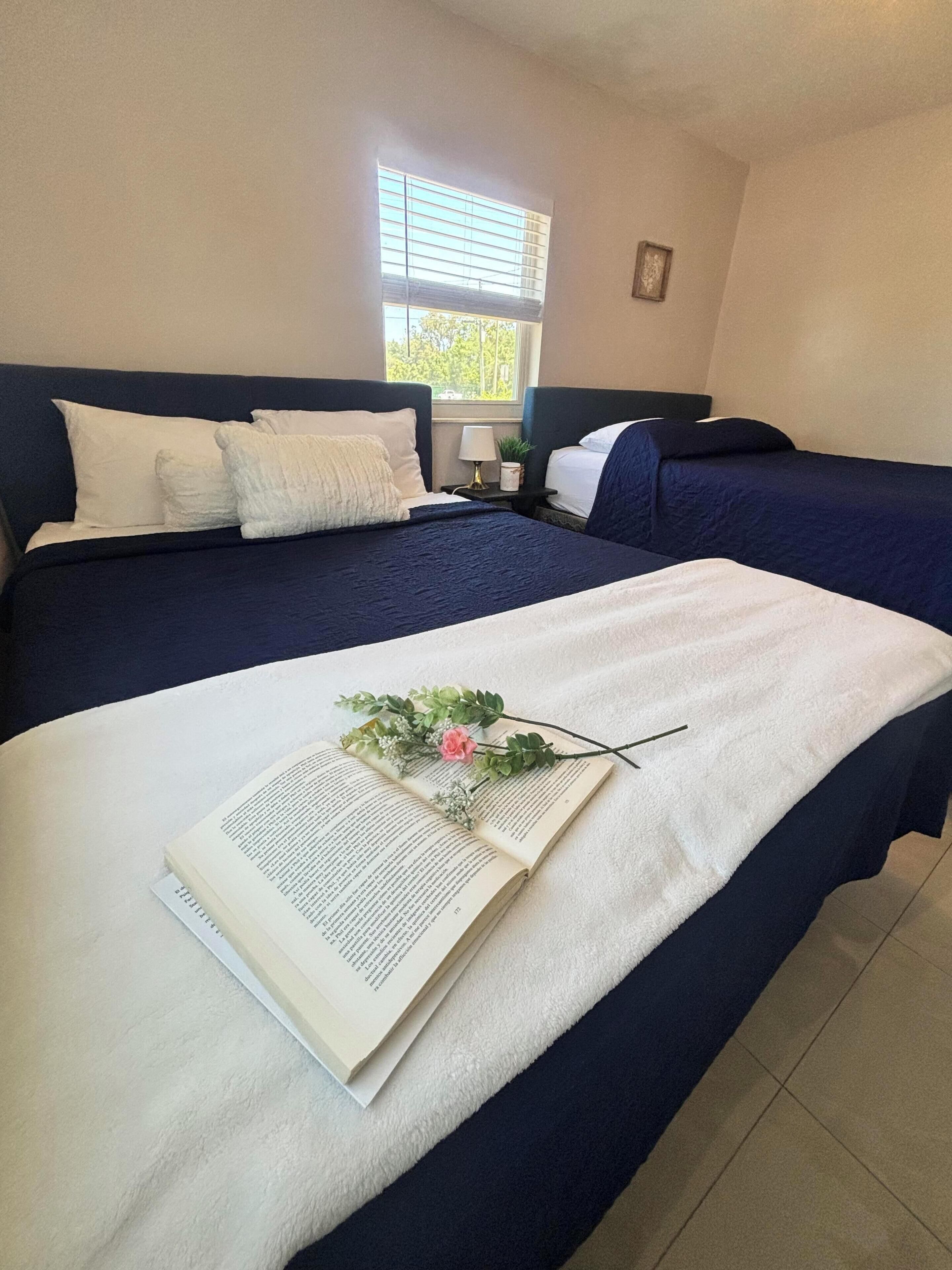 A hotel room with two beds, a window with blinds, a bedside table with a lamp, and a book with a flower arrangement on the bed.