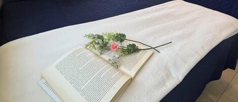 A hotel room with two beds, a window with blinds, a bedside table with a lamp, and a book with a flower arrangement on the bed.