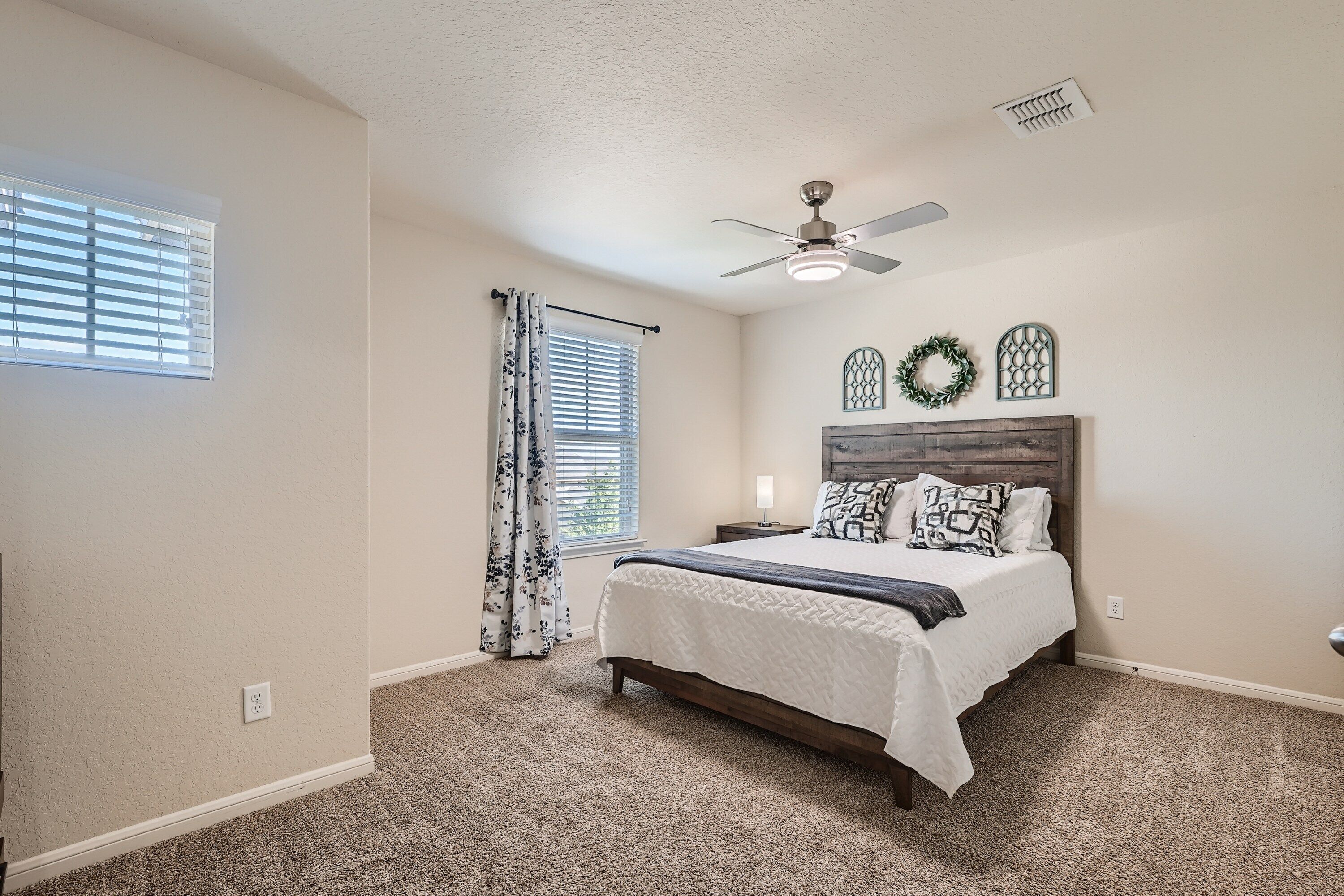 A bedroom with a bed, ceiling fan, window with blinds, and decorative wreaths on the wall.