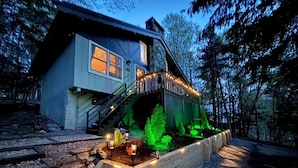 A house with a staircase leading to a deck, surrounded by a garden with illuminated plants. - Storybook Chalet/Watch Movies Under the Stars/NEW Hot Tub/Hiking/Biking/Concerts (Beech Mountain)