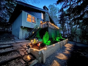 A house with a staircase leading to a deck, surrounded by a garden with illuminated plants.