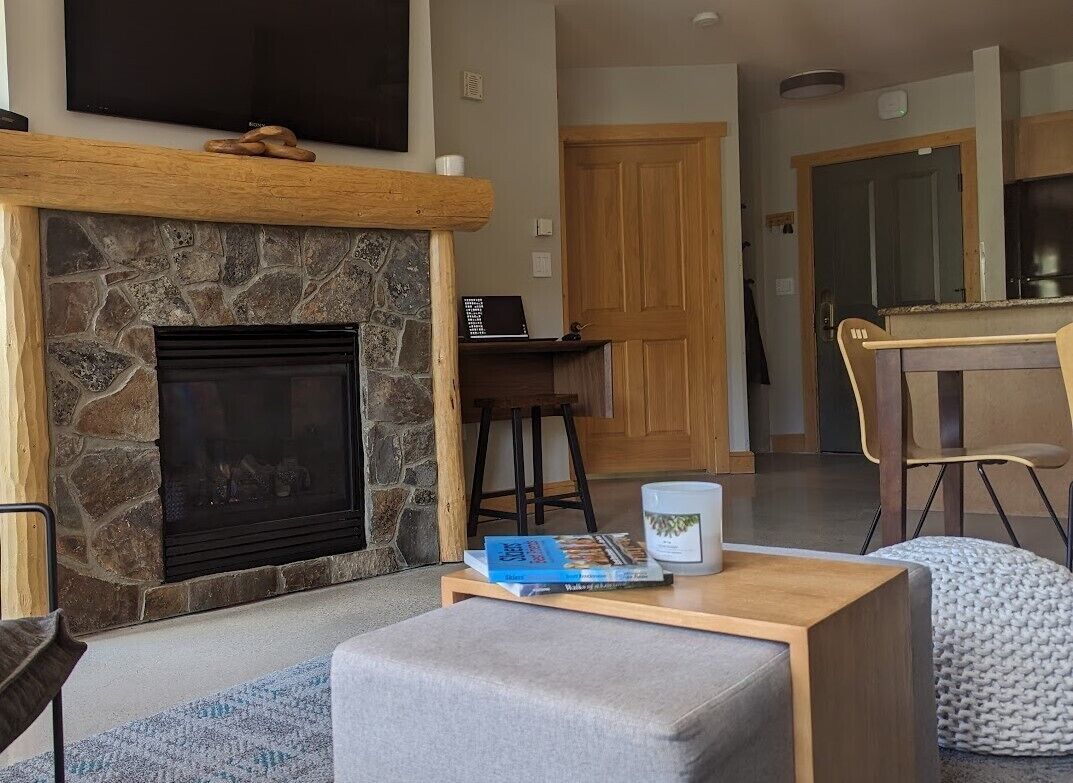 A living room with a stone fireplace, a flat-screen TV mounted above it, a wooden coffee table with books and a candle, and a cozy seating area with a cushioned bench and a knitted pouf.