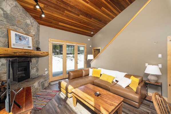 A cozy living room with a stone fireplace, wooden ceiling, and a large window overlooking a snowy landscape.