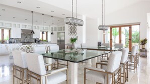 A spacious dining area with a glass table surrounded by upholstered chairs, a modern kitchen in the background, and large windows offering natural light. -  Gorgeous estate in Puerto Los Cabos golf and beach community (San José del Cabo)