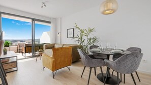 A modern living room with a sofa, chairs, a dining table set for four, and a large window offering a view.