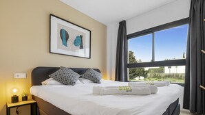 Een hotelkamer met een groot bed, een kunstwerk erboven, een nachtkastje met een lamp en uitzicht op het landschap door een groot raam.