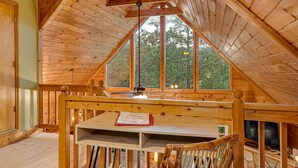 Ein gemütliches Holzhaus mit geneigter Decke, einem großen Fenster mit Blick auf Bäume, einer hölzernen Bank und einem Tisch mit einer Willkommensschild.