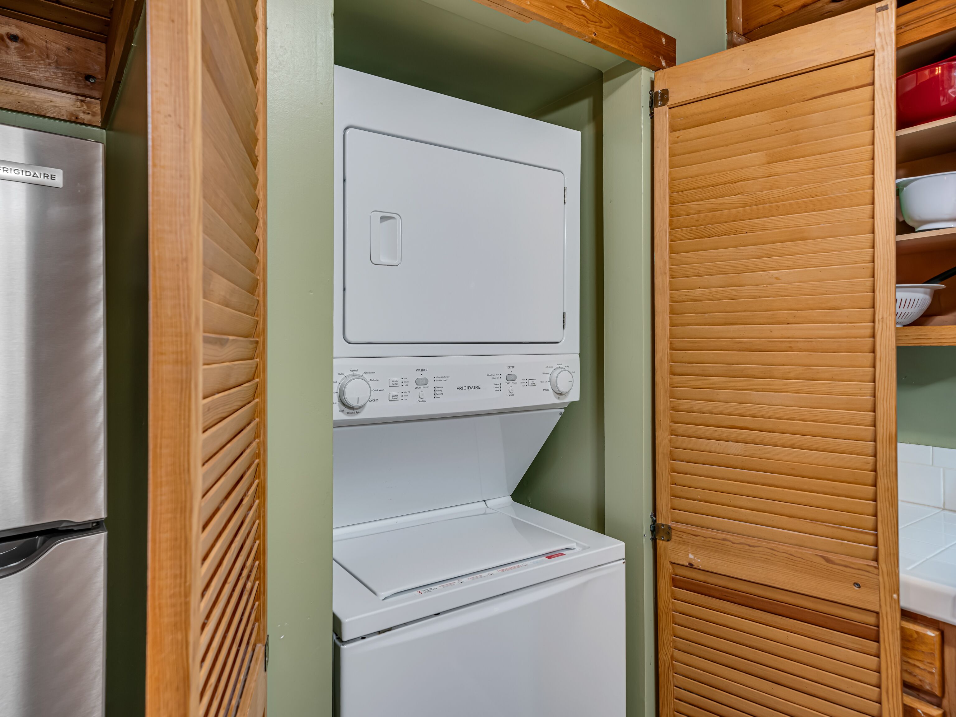 Une petite buanderie équipée d’un lave-linge et d’un sèche-linge Frigidaire, de stores en bois et d’une étagère où sont rangés de la vaisselle.