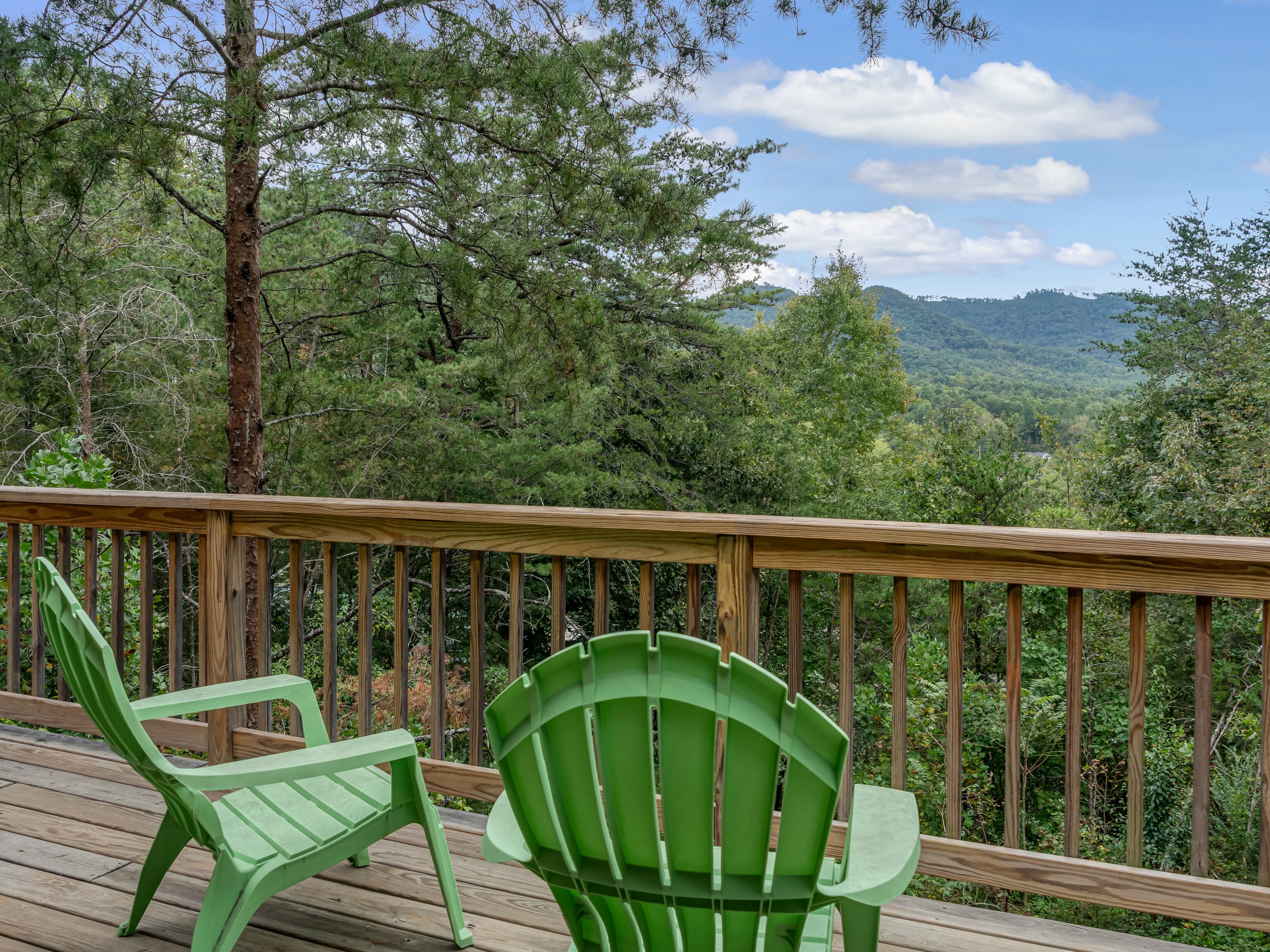A wooden deck with two green Adirondack chairs overlooking a forested landscape.