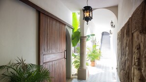 Un pasillo con puerta de madera, plantas en macetas, suelo de baldosas y pared de piedra con esculturas.