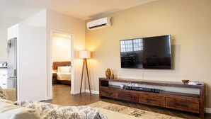 Una sala de estar moderna con un televisor de pantalla plana, un mueble de entretenimiento de madera, un sofá y un área de comedor visible a través de un vano abierto.