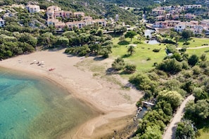 Luftaufnahme einer Küstenregion mit Sandstrand, klarem Wasser und umgebender Bepflanzung.
