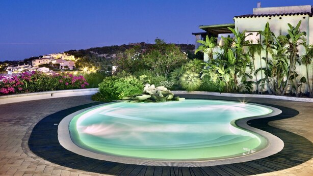 Une piscine à bordures ondulées, entourée de plantes tropicales, offrant une vue sur une ville perchée sur une colline au crépuscule.