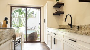 A modern kitchen with white cabinets, a granite countertop, and a black faucet. There is a sliding glass door leading to a patio with a wicker chair and a plant. - Casa Ixchel | A Modern Mexican Oasis | Private Pool (San Miguel de Cozumel)