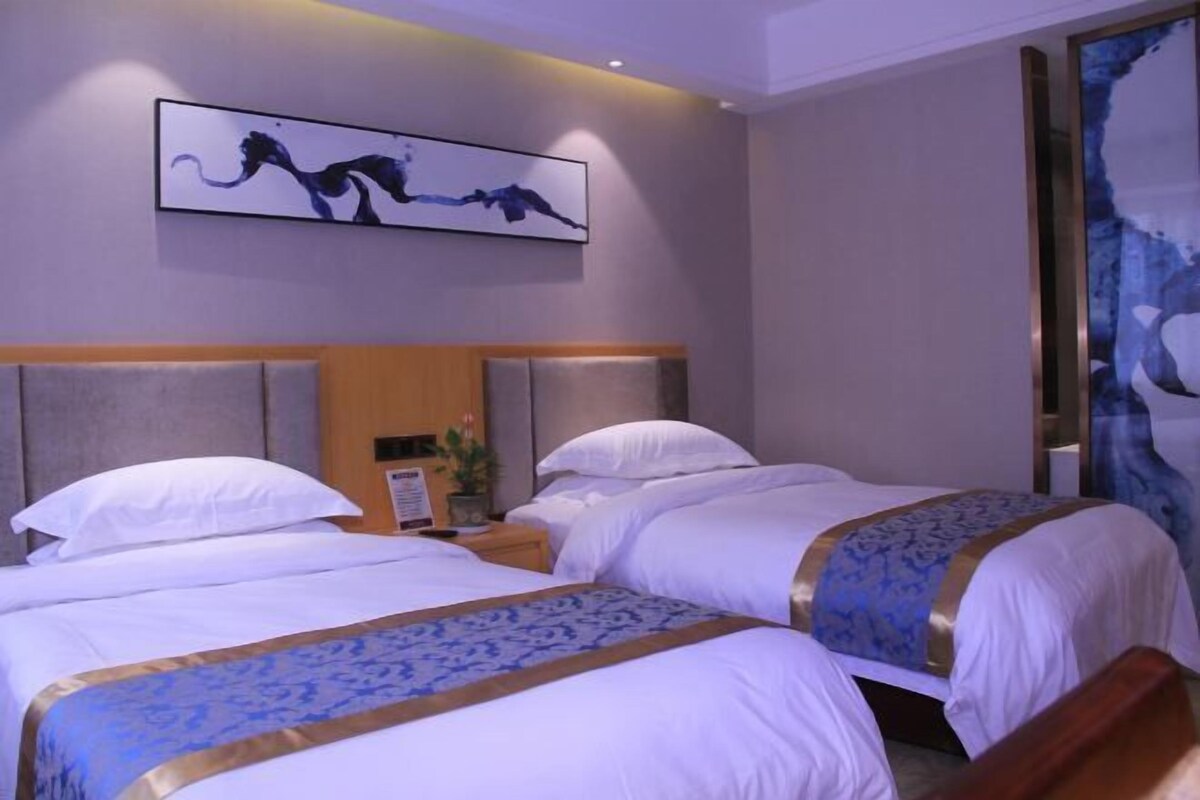 Hotellihuone, jossa on kaksi sänkyä, sängynpääty, seinälle ripustettu taideteos ja suuri ikkuna.