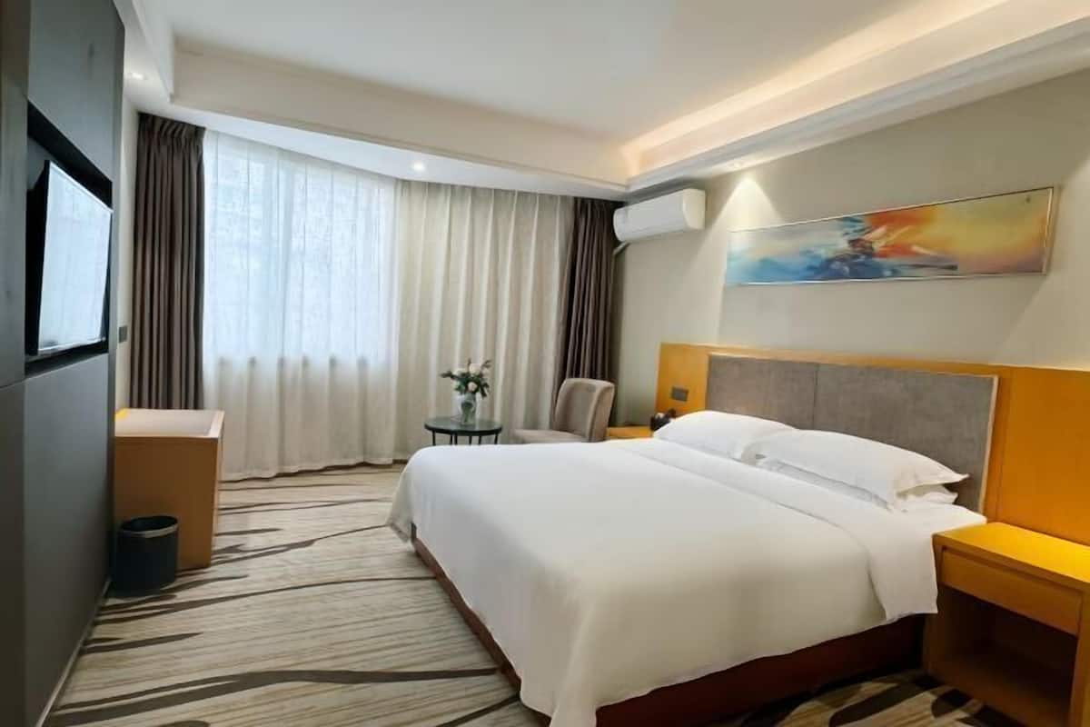 A hotel room with a large bed, a desk, a chair, a small table with a vase, and a painting on the wall.