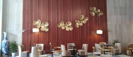 A modern hotel lobby with a red panel wall, a large abstract gold wall art piece, and a seating area with round tables and chairs.