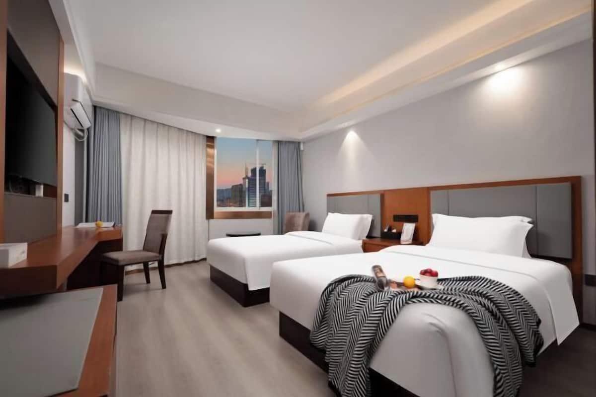 A hotel room with two beds, a desk, a chair, a TV, and a window with a cityscape view.