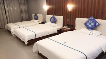 Habitación de hotel con tres camas individuales, cada una con una almohada azul y una colcha blanca. Hay dos mesitas de noche con lámparas, y las paredes están revestidas de paneles de madera.