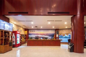 A reception area with a wooden reception desk, a red vending machine, and a wall with a scenic mural. - Gujiang Hotel (Dahua)