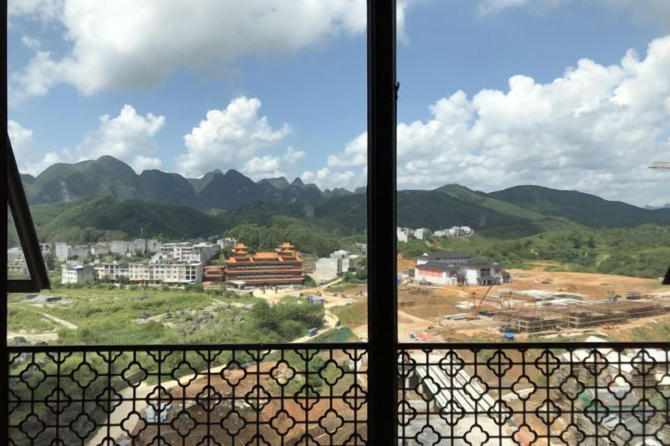 Une vue à travers une fenêtre ornée d’un motif géométrique, donnant sur un paysage montagneux avec des bâtiments et des chantiers de construction.