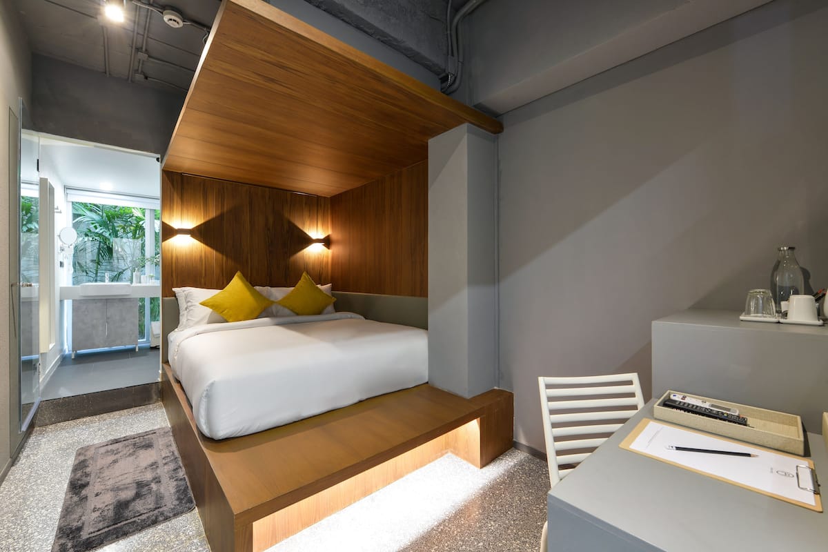 A modern bedroom with a wooden headboard, a bed with white bedding and yellow pillows, a desk with a chair, and a view of a balcony.