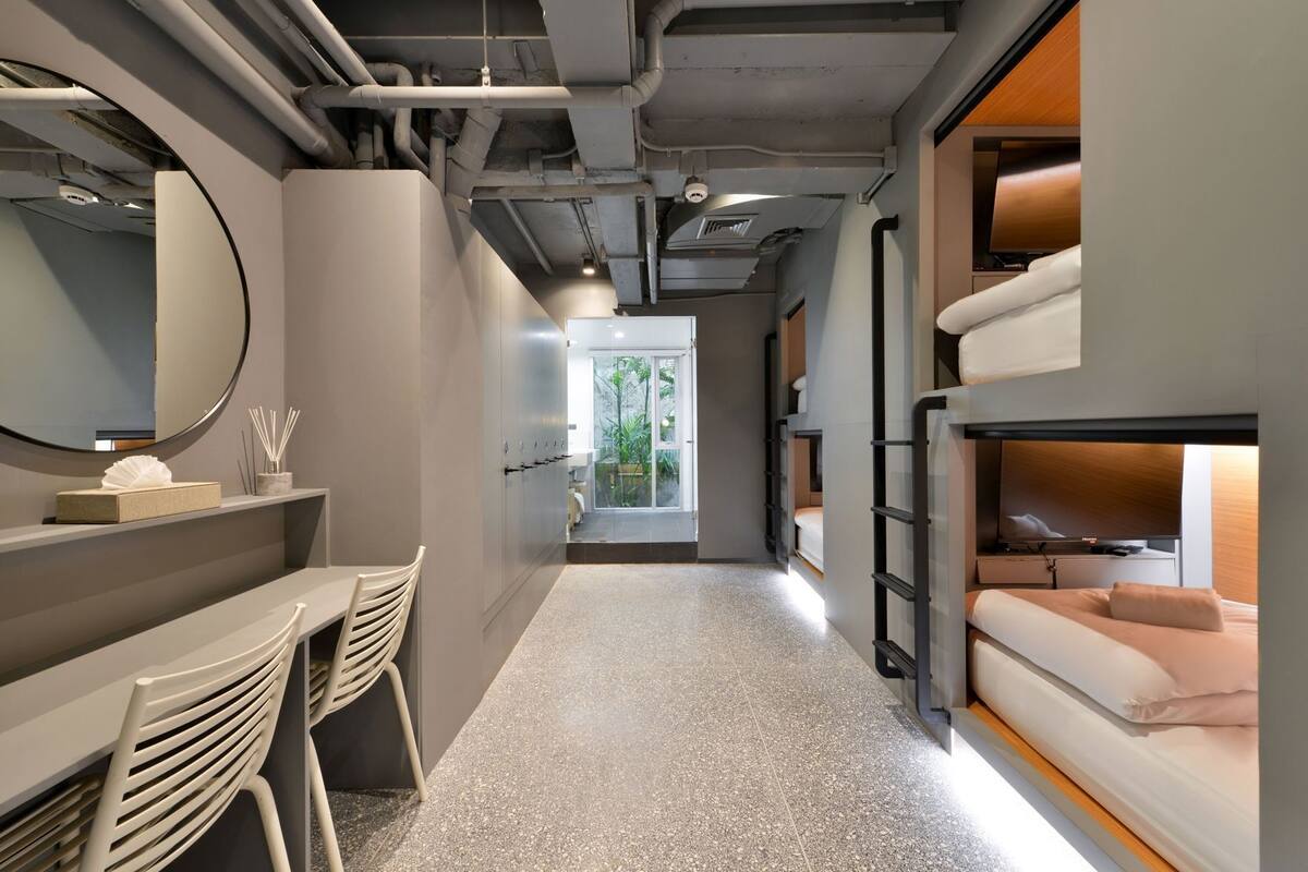 A modern hotel room with bunk beds, a desk with chairs, a large mirror, and a view of greenery outside.