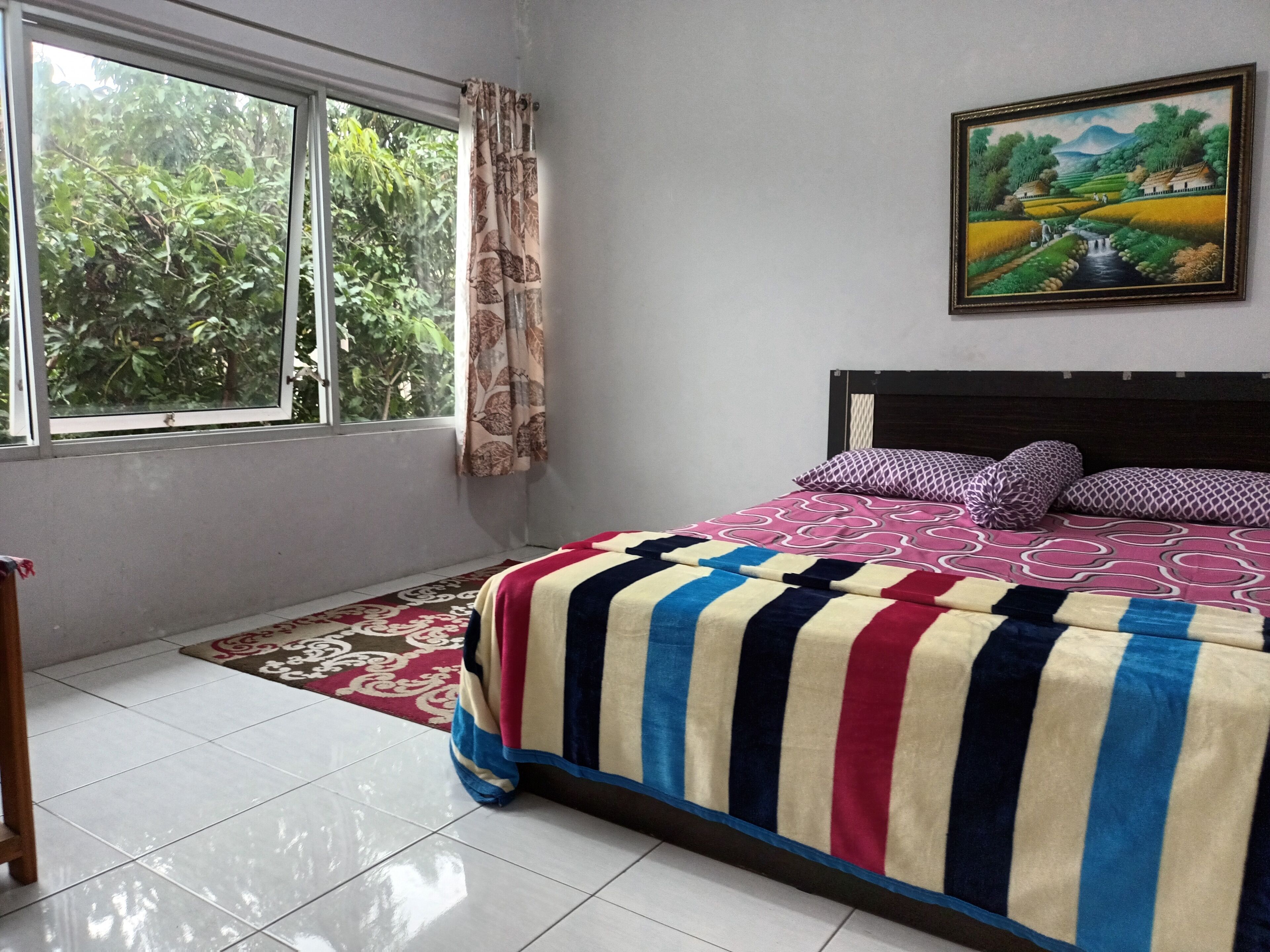 A bedroom with a large bed, a window with curtains, a painting on the wall, and a tiled floor.