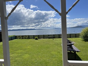 A view from a covered area overlooking a lake with a wooden fence and picnic tables. - Daydream House, Peaceful, sunrise/set views across the lake (Rotorua)