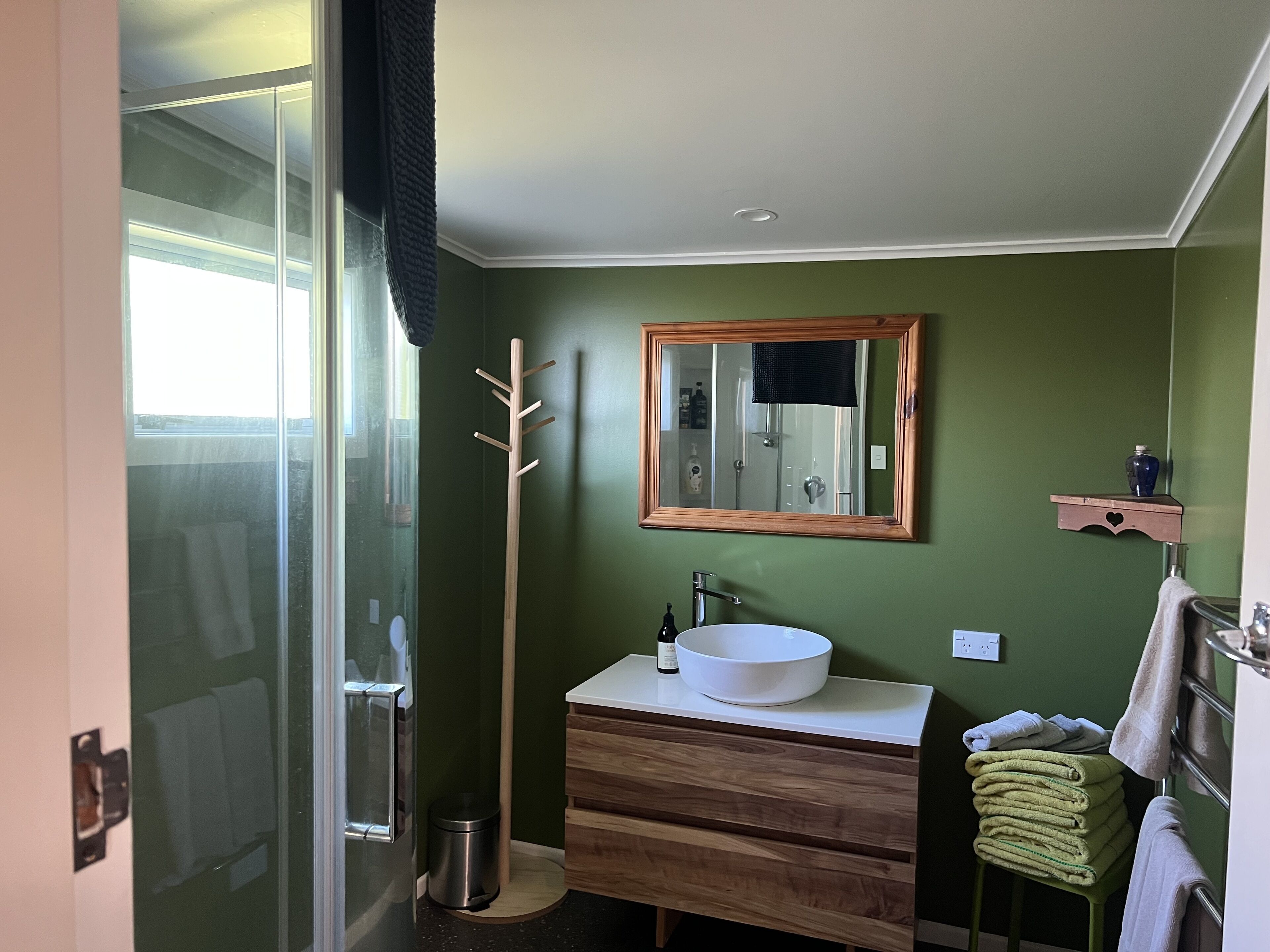 A bathroom with a glass shower door, a wooden coat rack, a sink with a white basin, and a mirror with a wooden frame.