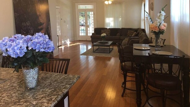 A spacious living room with a dark brown sofa, a coffee table, and a dining area with a table and chairs. There is a large window with curtains and a ceiling fan.