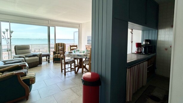 A room with a view of the sea, a dining area, and a kitchen with a red trash bin.