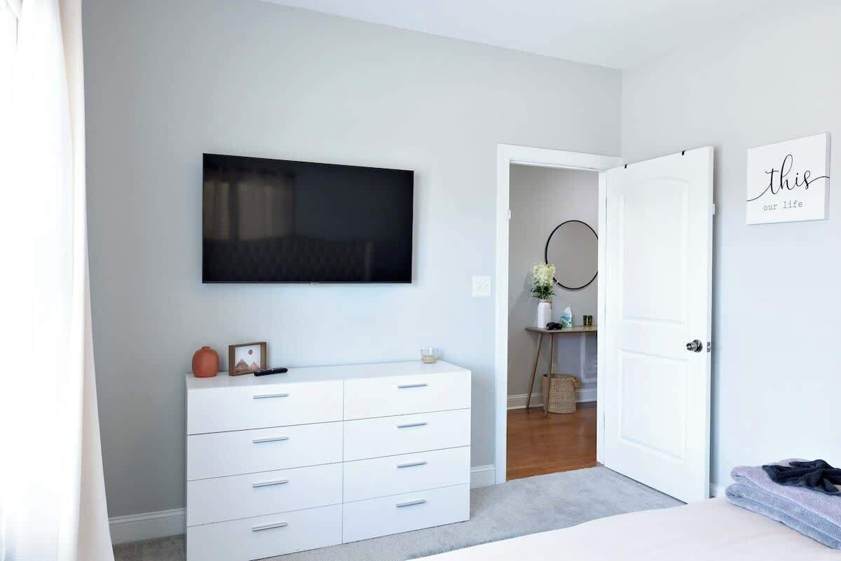Ein Schlafzimmer mit einem Bett, einer weißen Kommode, einem flachbildfernseher an der Wand und einer Tür zu einem weiteren Zimmer.