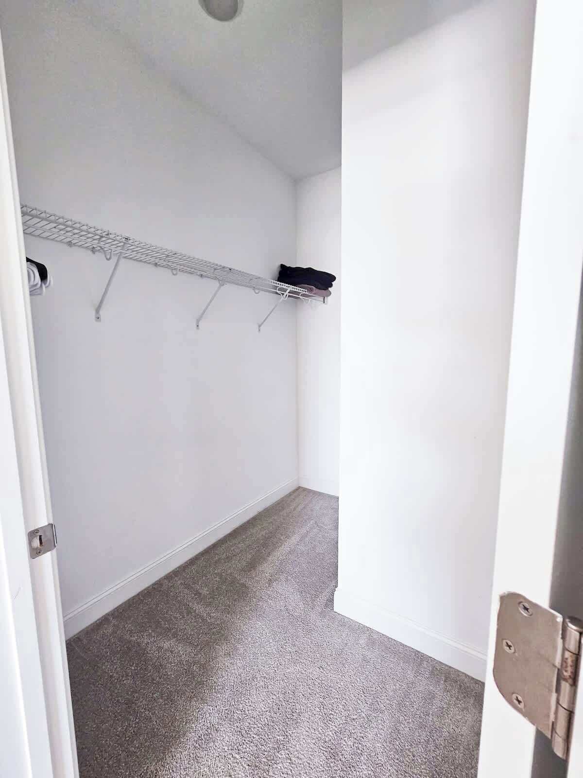 Una habitación pequeña y vacía, con alfombra gris, paredes blancas y un estante de metal con ropa colgada.