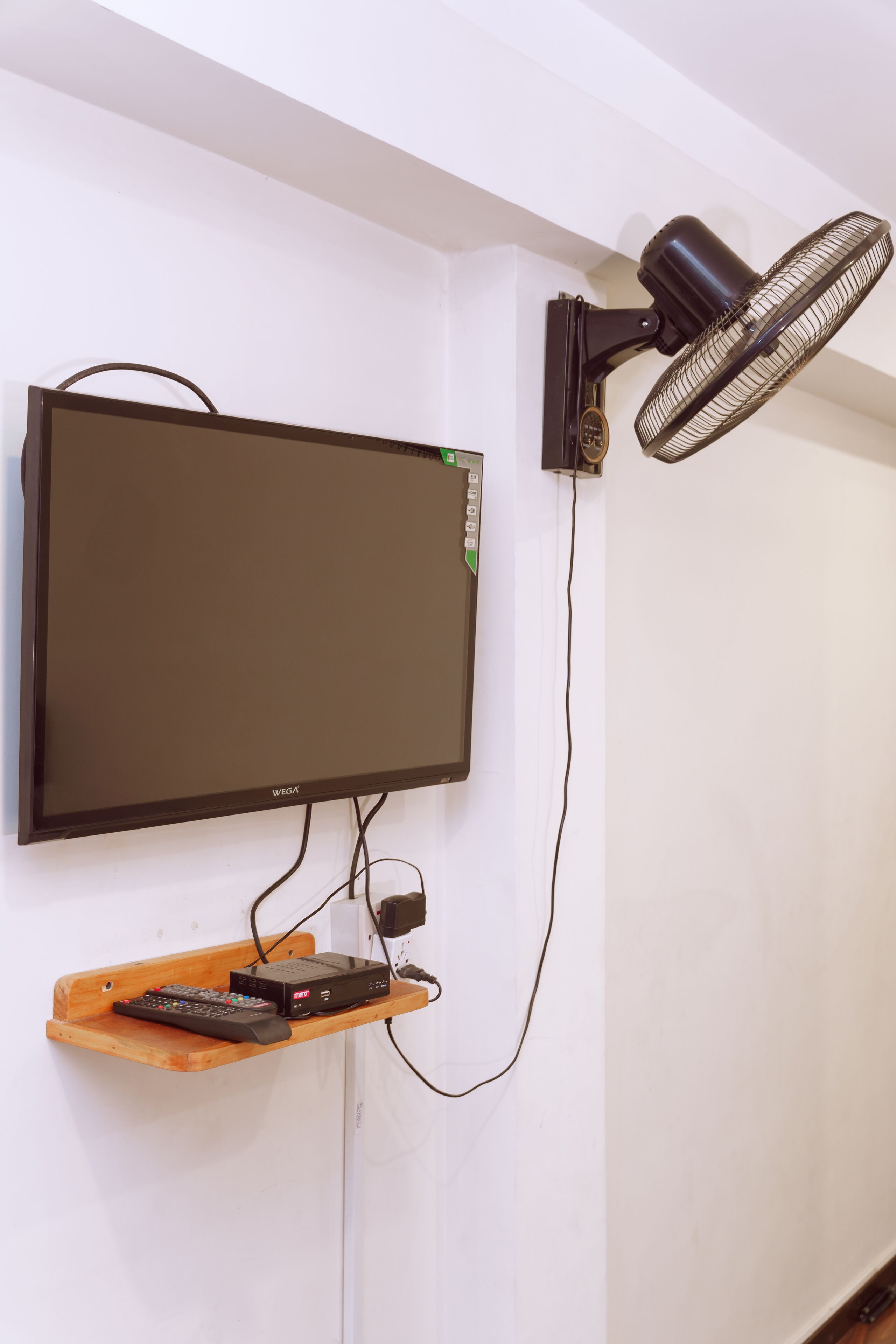 Un televisor de pared con control remoto sobre un estante de madera, un ventilador negro y una regleta de enchufes.