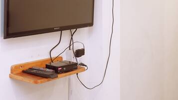Un televisor de pared con control remoto sobre un estante de madera, un ventilador negro y una regleta de enchufes.